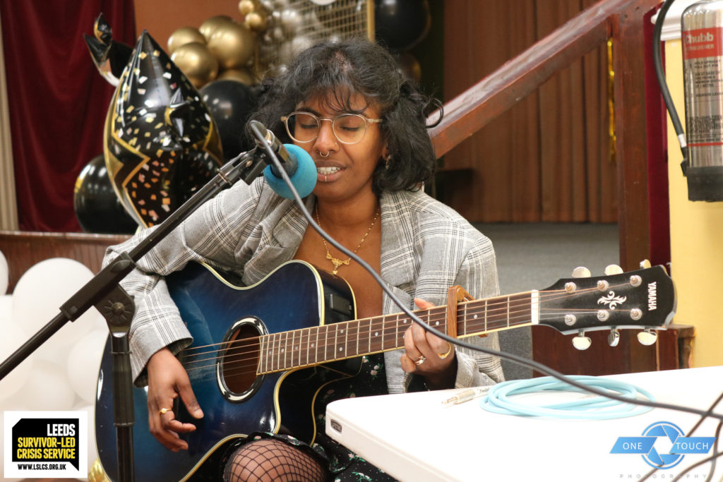 A dark skinned feminine person with glasses playing an acoustic guitar